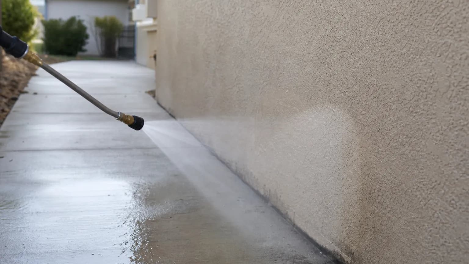 Low pressure fan of cleaning solution being sprayed from a wand onto a beige stucco wall, with a clean concrete walkway running along the base of the wall